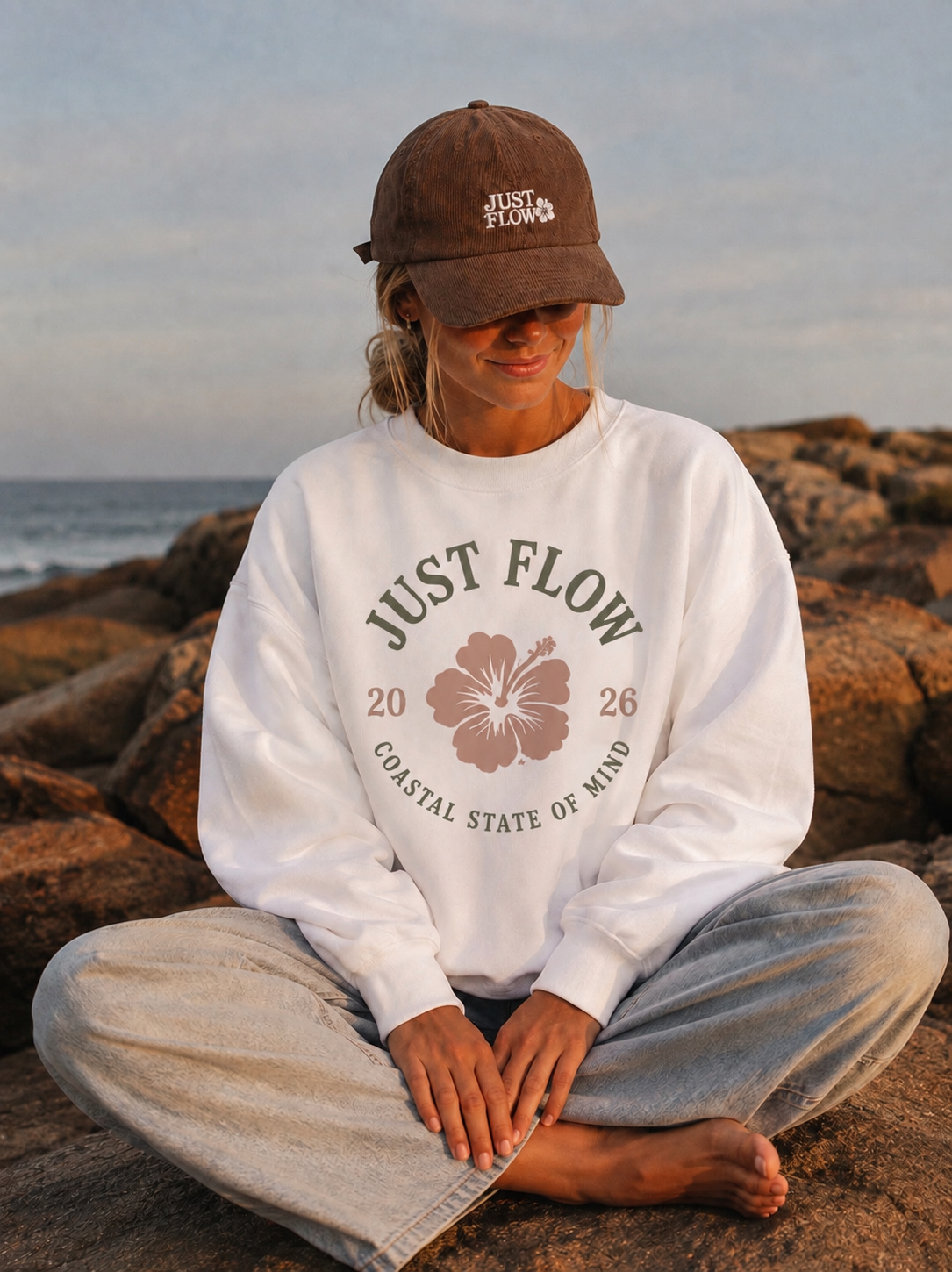 Hibiscus Badge Crew Sweatshirt in white, seated on coastal rocks